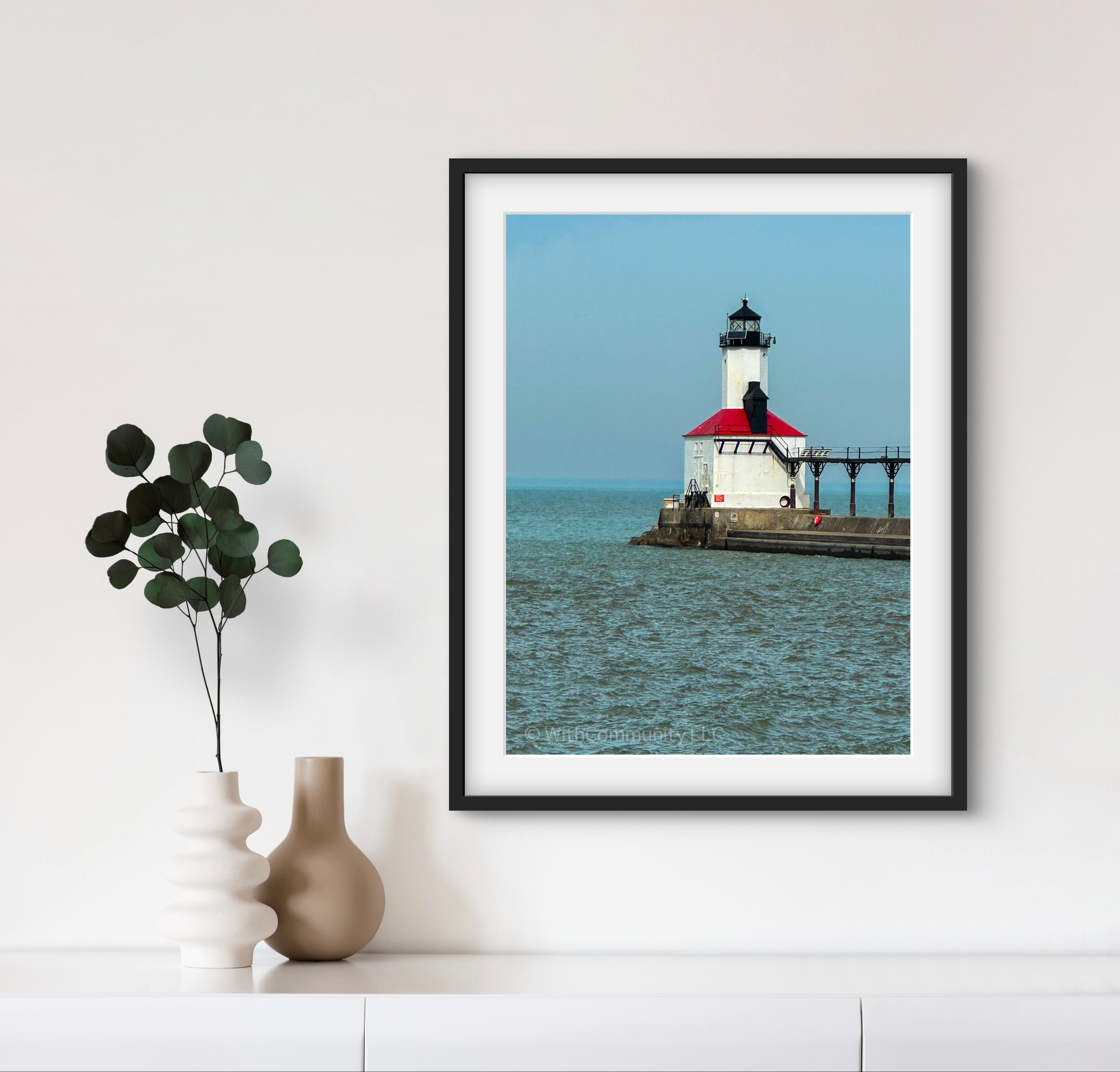Framed picture of a lighthouse on a wall with a vase and plant below.