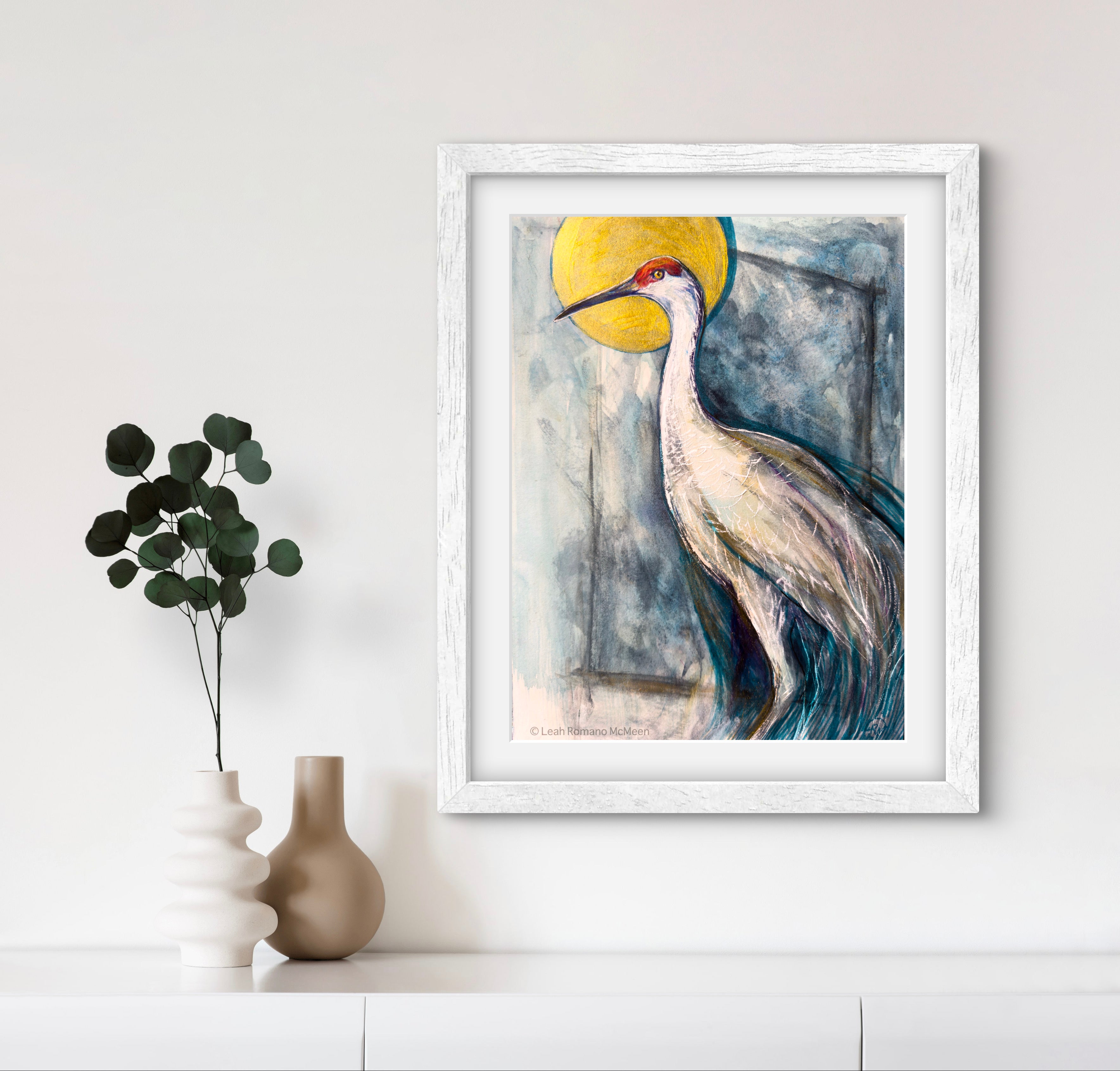 Framed artwork of a crane bird with a yellow sun on a white wall.