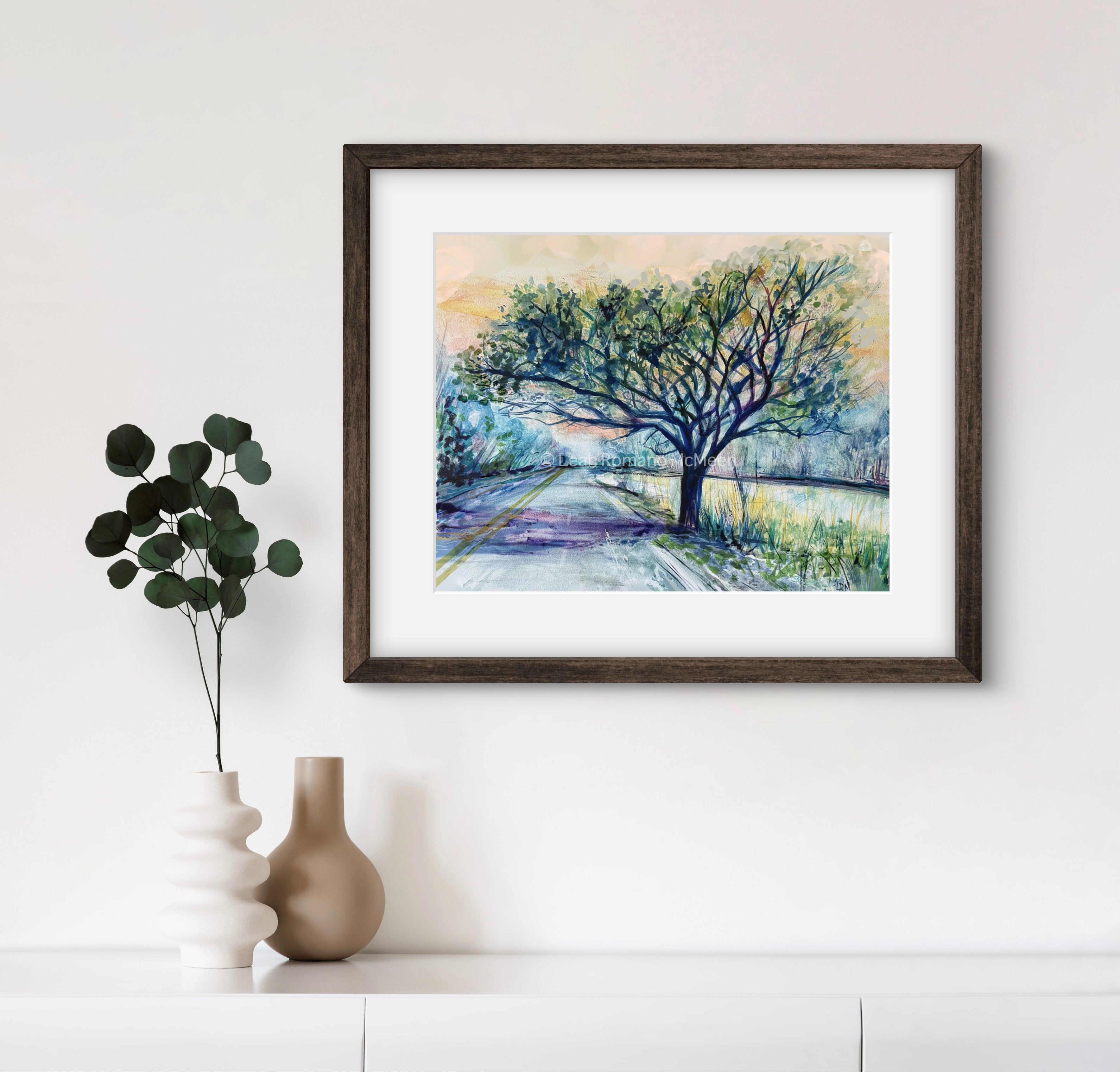 Framed artwork of a tree on a path with a vase and plant on a shelf below.