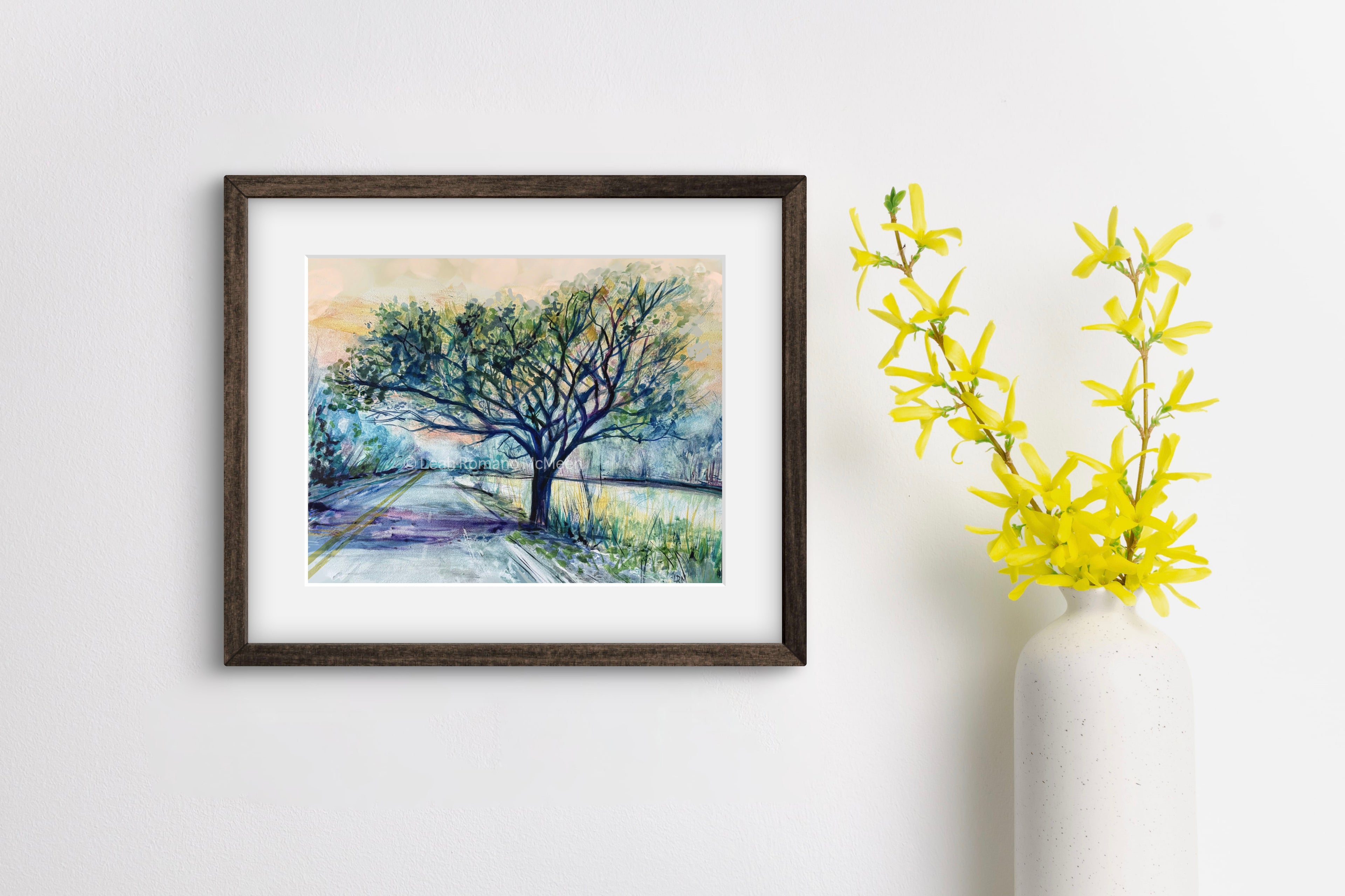 Framed artwork of a landscape with a tree on a white wall next to a yellow plant.